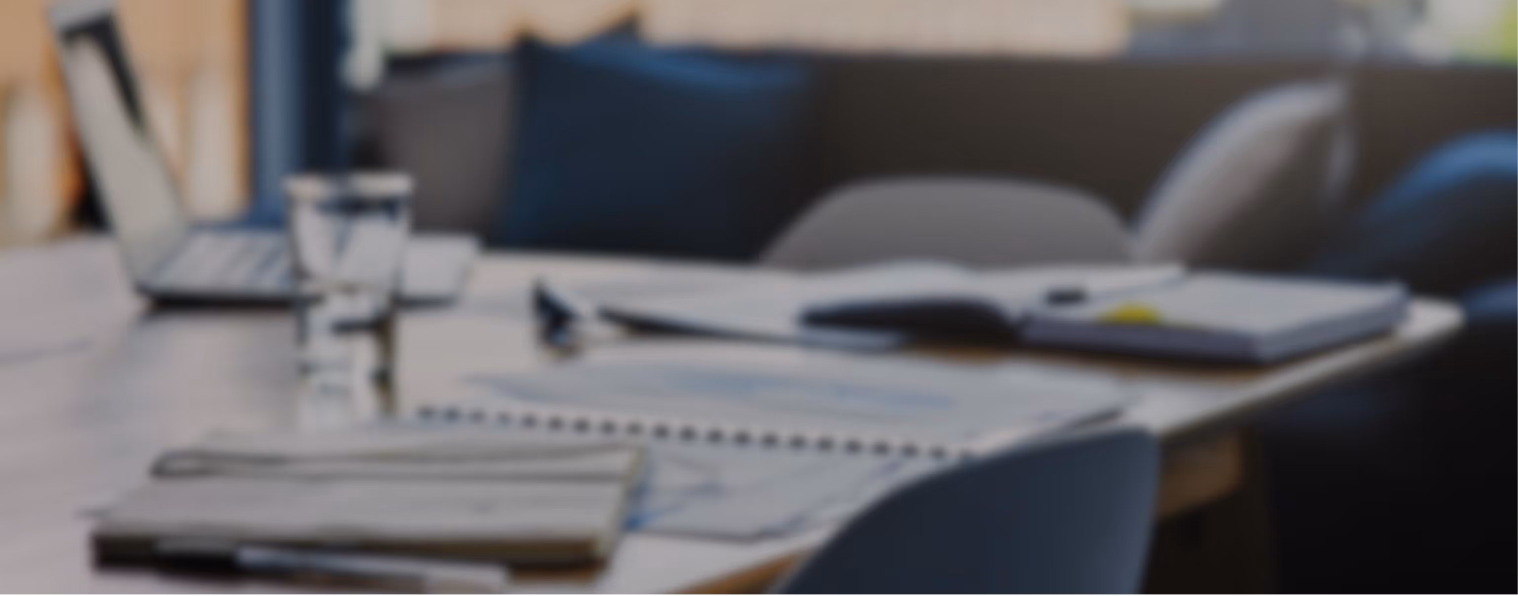 Blurred image of a workspace with a laptop, open notebook, glass of water, and documents on a wooden table.