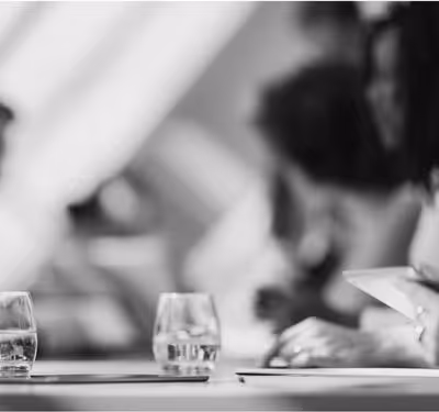 Blurred grayscale image of people sitting at a table with glasses of water and a hand holding a phone.