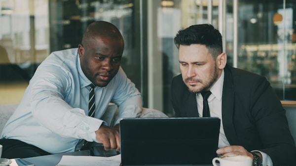 Two business colleagues looking at laptop computer and discussing their startup plan in modern office with glass walls. Bearded businessman and his partner sitting at table and talking future deals
