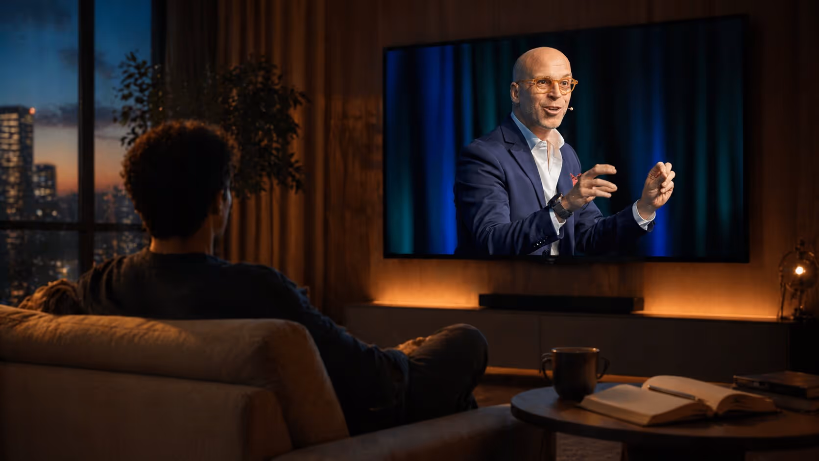 Person sitting on a couch watching a bald man in a suit speaking on a large TV screen in a cozy living room at dusk.
