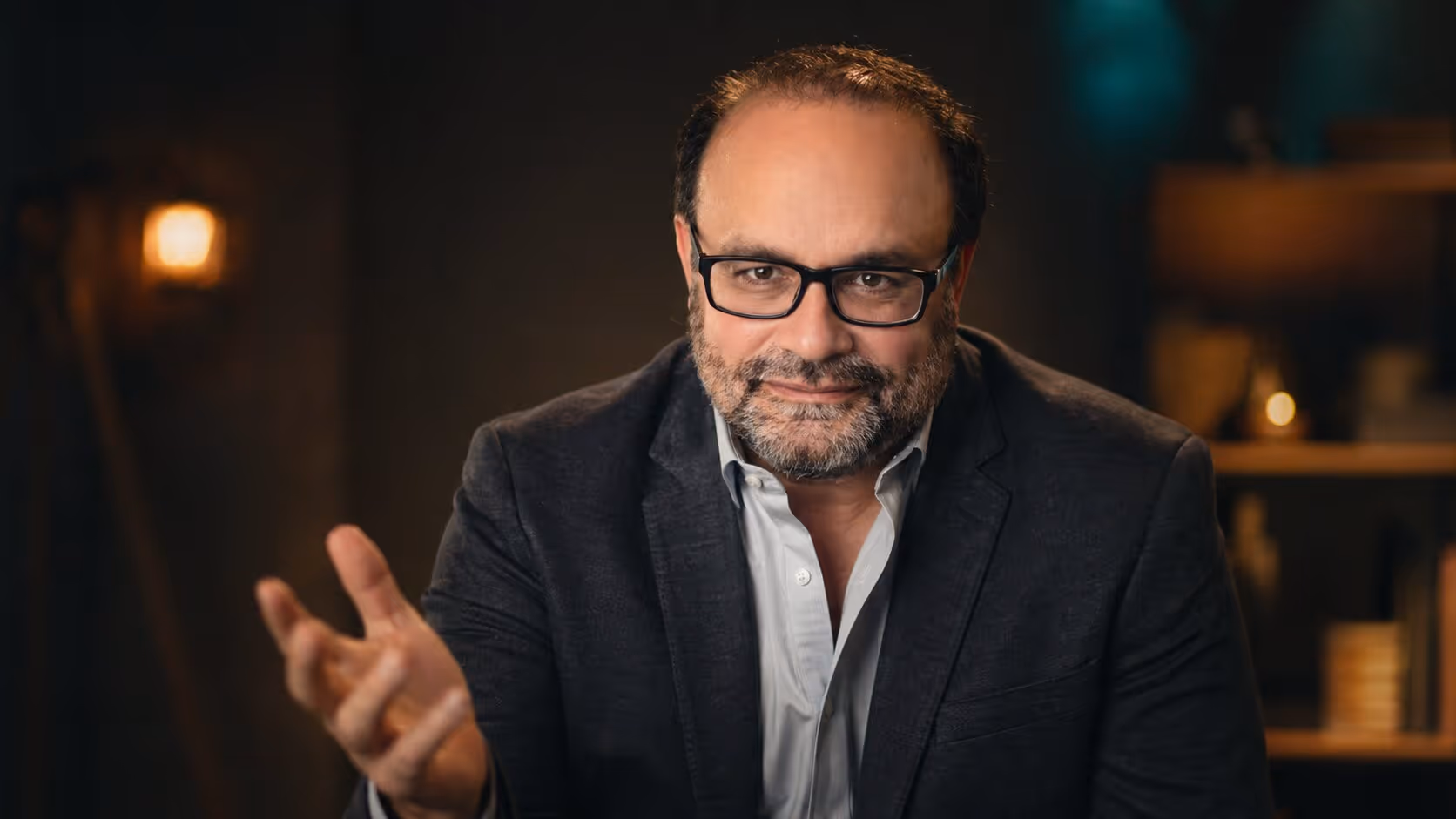 Middle-aged man with glasses and beard wearing a dark blazer and light shirt, gesturing with one hand.