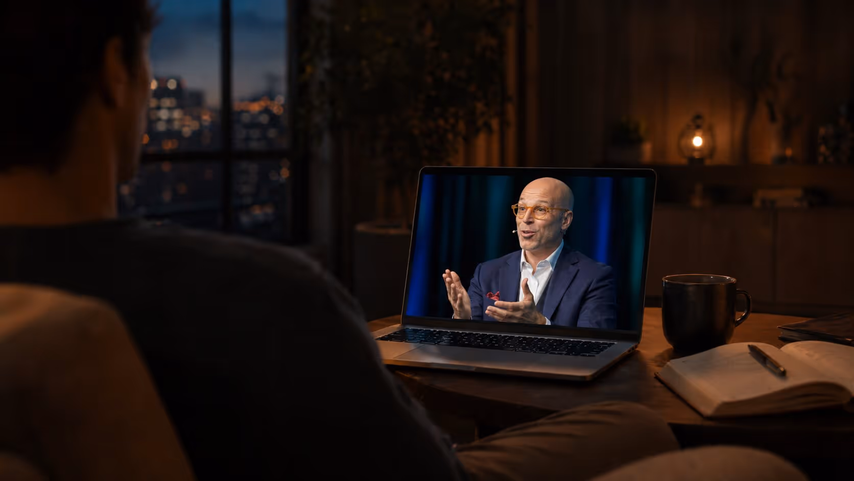 Person watching a bald man in glasses speaking on a laptop screen during a video call in a dimly lit room.