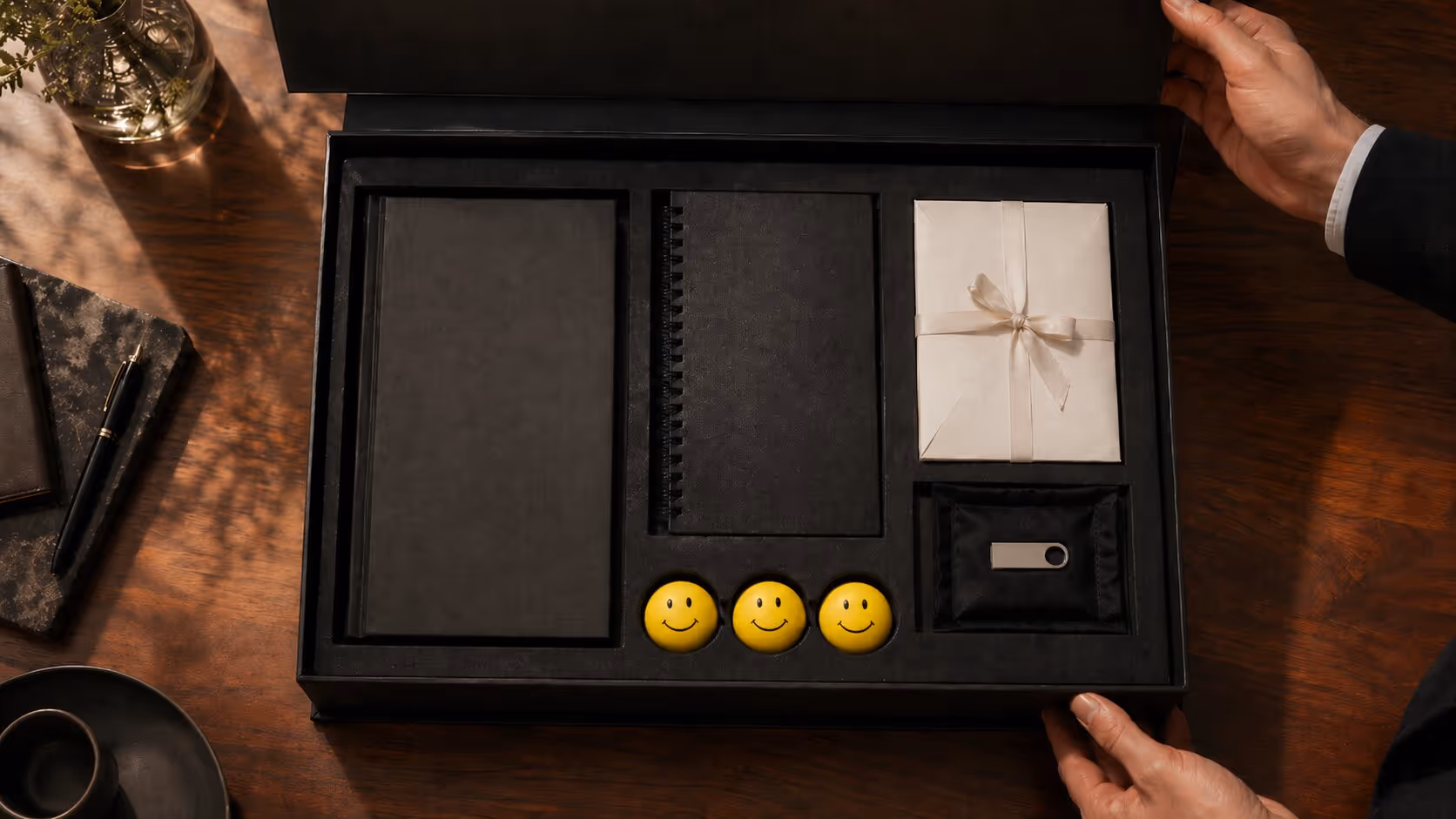 Unboxed gift set with black notebooks, three yellow smiley face stress balls, a wrapped envelope, and a USB flash drive on a wooden table.