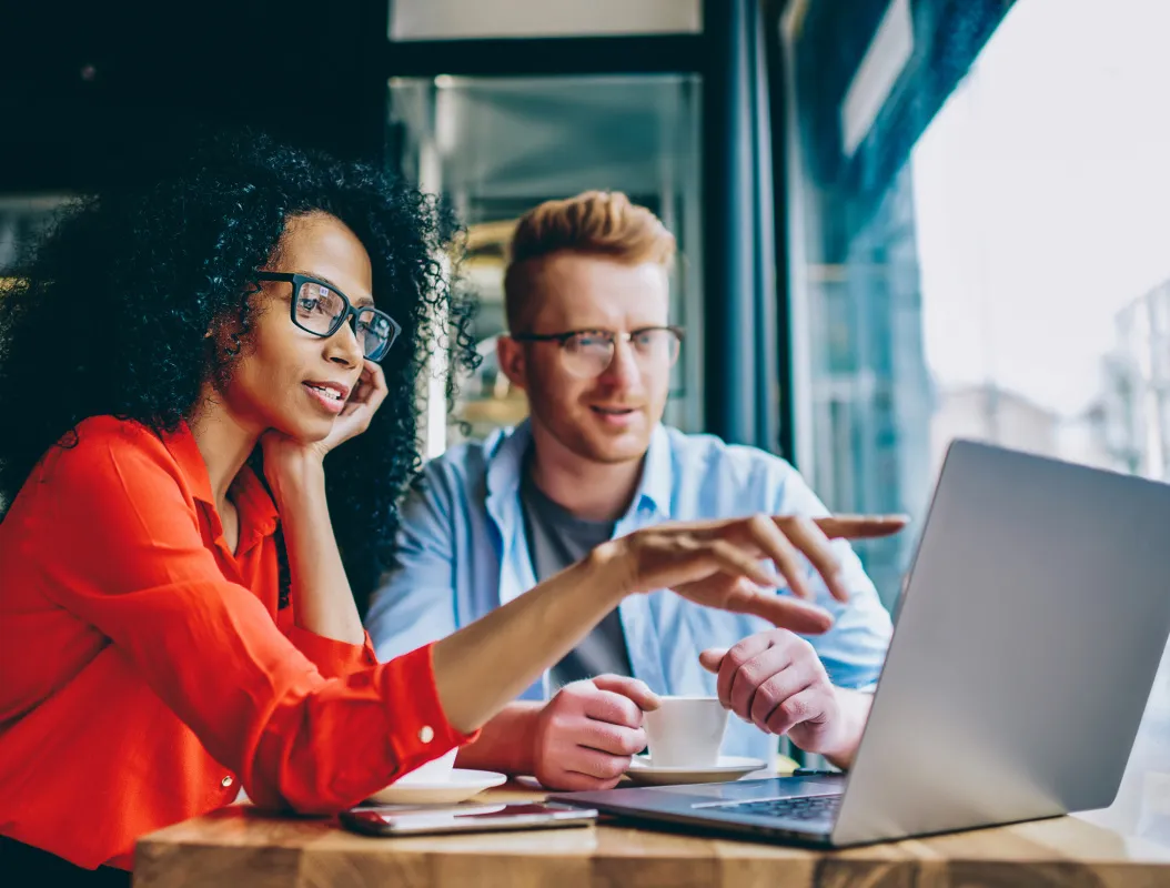 Two colleagues wearing glasses, sitting at a table by a window, looking at a laptop while one points at the screen.