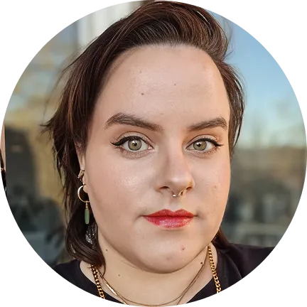 Close-up portrait of a person with short brown hair, green eyes, wearing red lipstick, a septum ring, and dangling earrings.