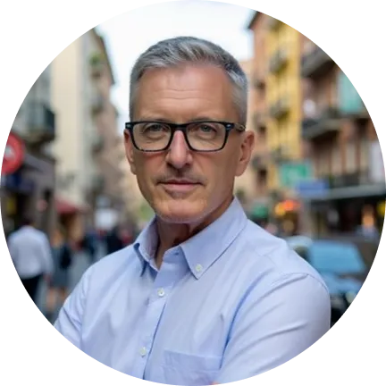 Middle-aged man with short gray hair and glasses wearing a light blue button-up shirt, standing outdoors on a city street.