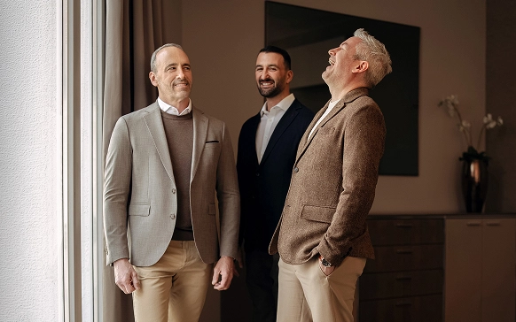 Three men in business casual attire standing indoors, with two smiling and one laughing while facing each other.