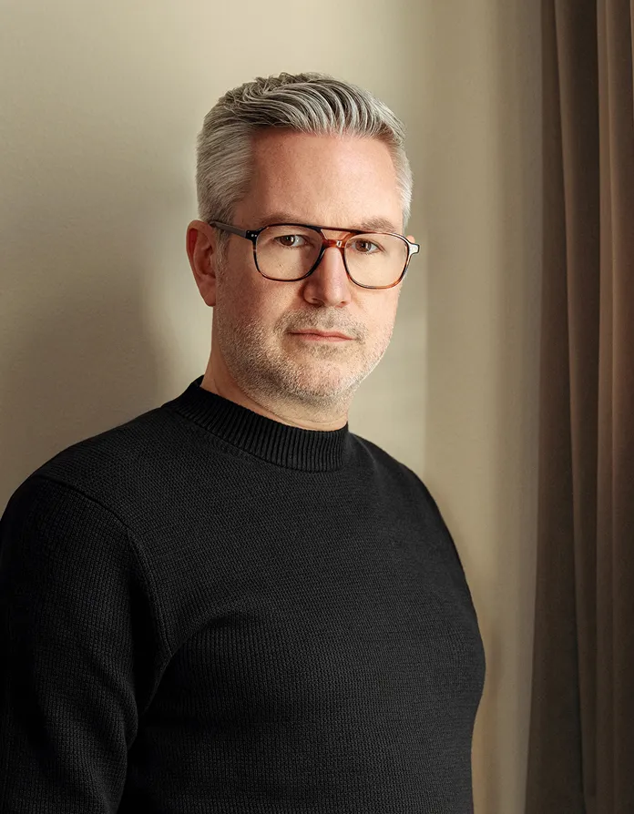 Middle-aged man with gray hair and glasses wearing a black sweater standing indoors against a beige wall and curtain.