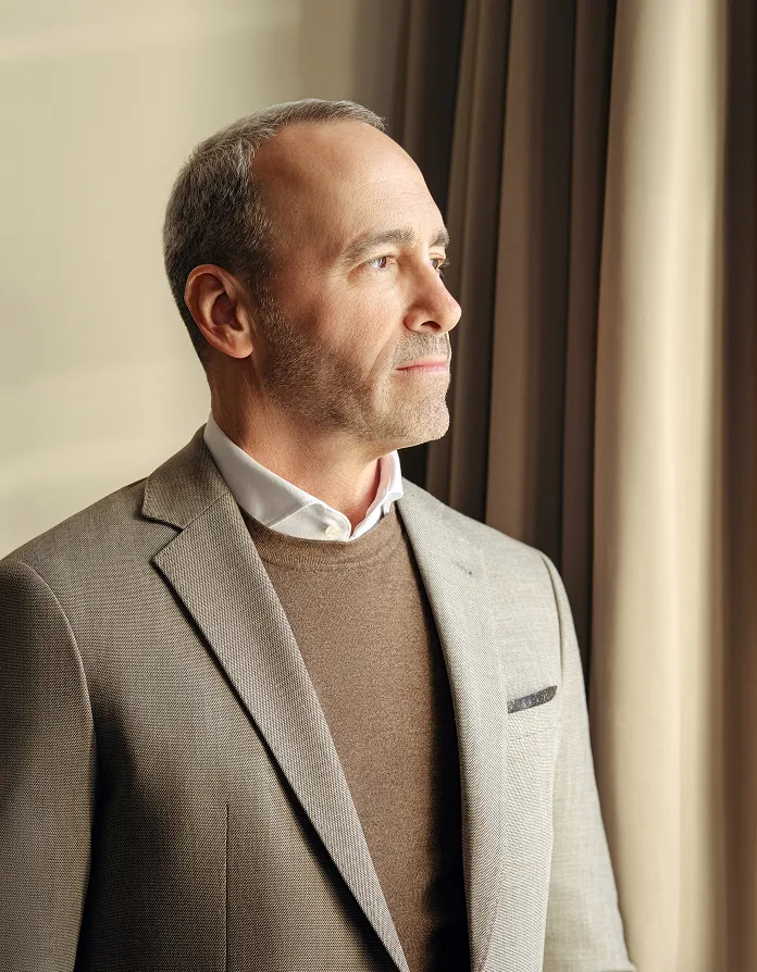 Middle-aged man with short hair and light stubble wearing a beige blazer, brown sweater, and white shirt against a neutral background.