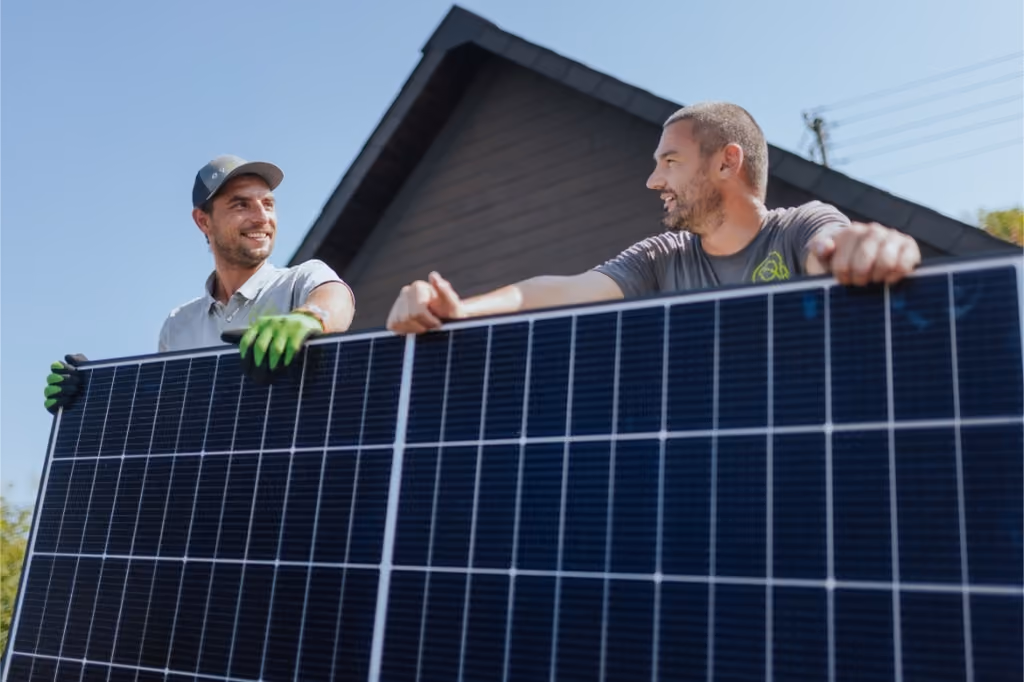 Zwei Männer tragen ein großes Solarmodul vor einem Haus mit Dach.