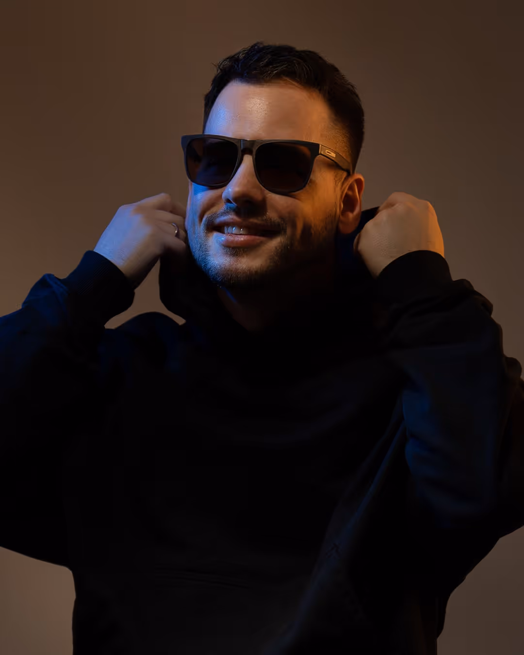 Man wearing sunglasses and a black hoodie, smiling while holding the hoodie collar with both hands against a dark background.
