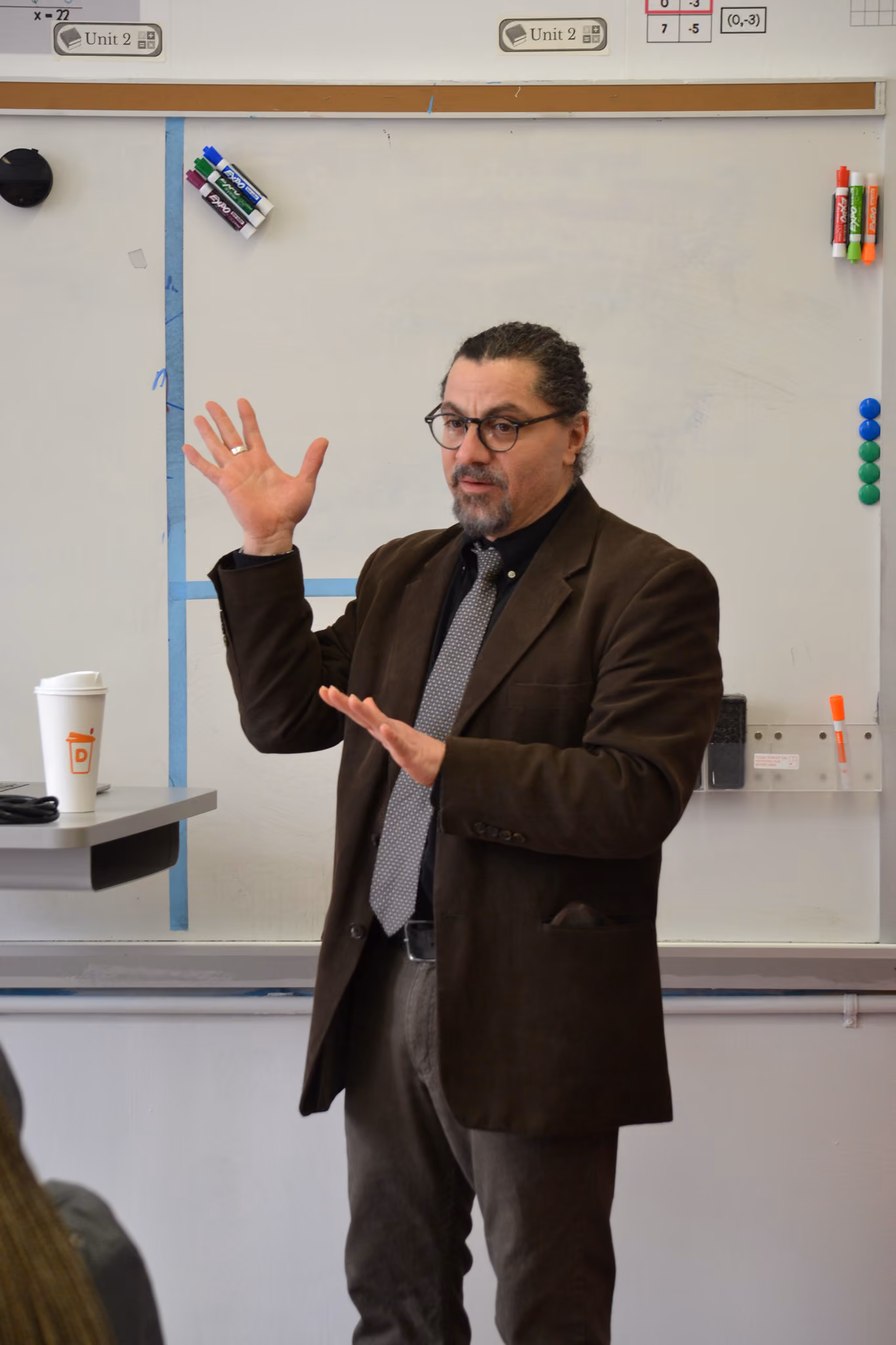 Teacher in suit gesturing while speaking in front of whiteboard