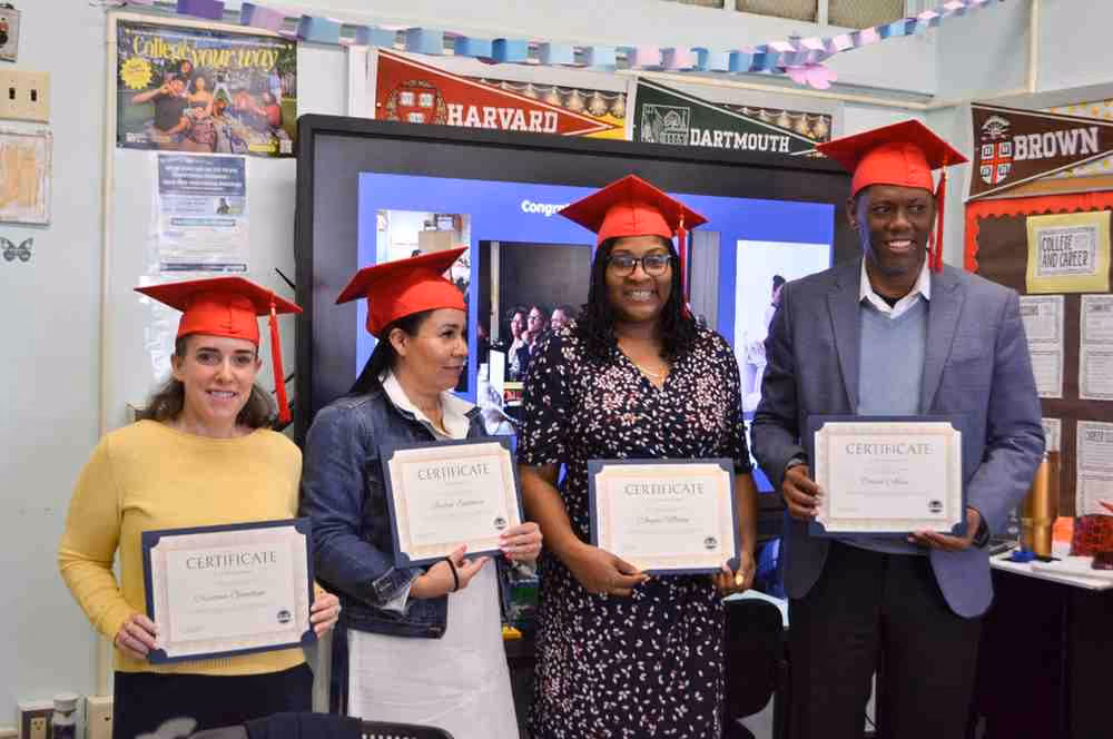 Four graduates in red caps holding certificates in decorated classroom