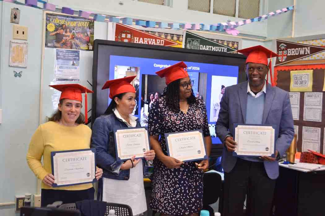 Four graduates in red caps holding certificates in decorated classroom