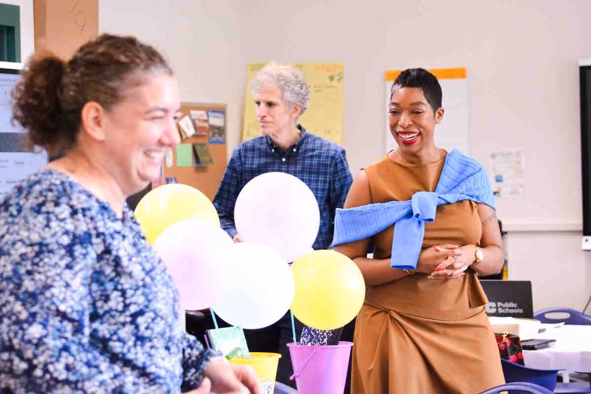 Three adults smiling during a school celebration with colorful balloons