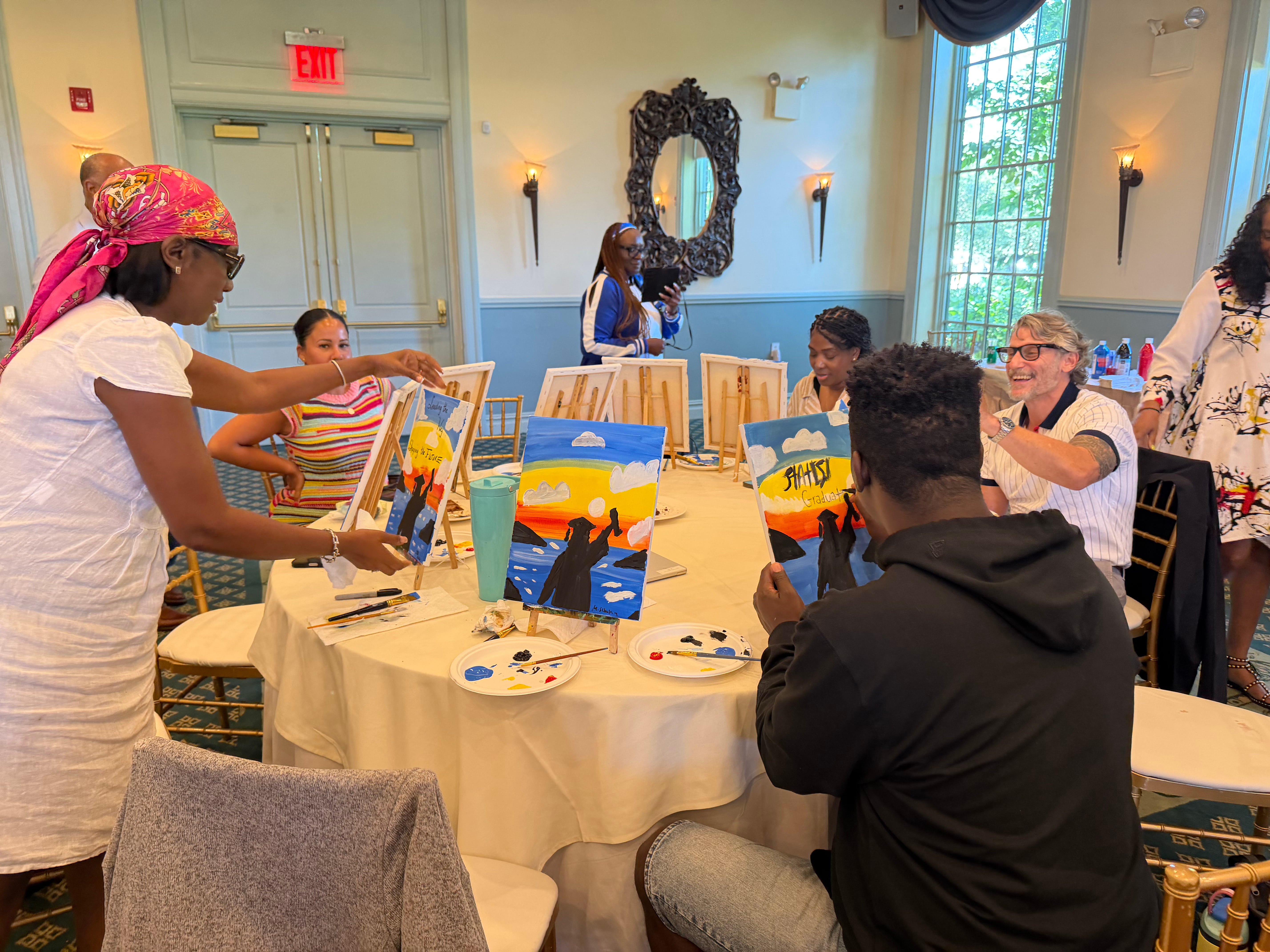 Students and adults participate in a group painting activity at round tables