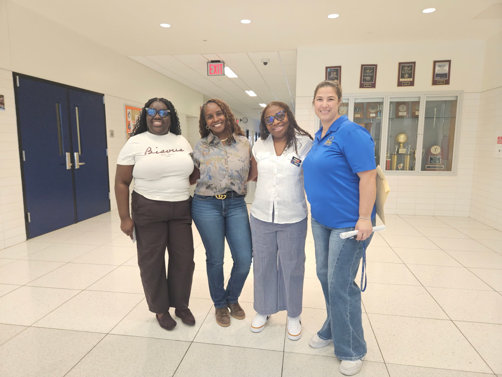 Four women posing together in a school hallway near trophy cases