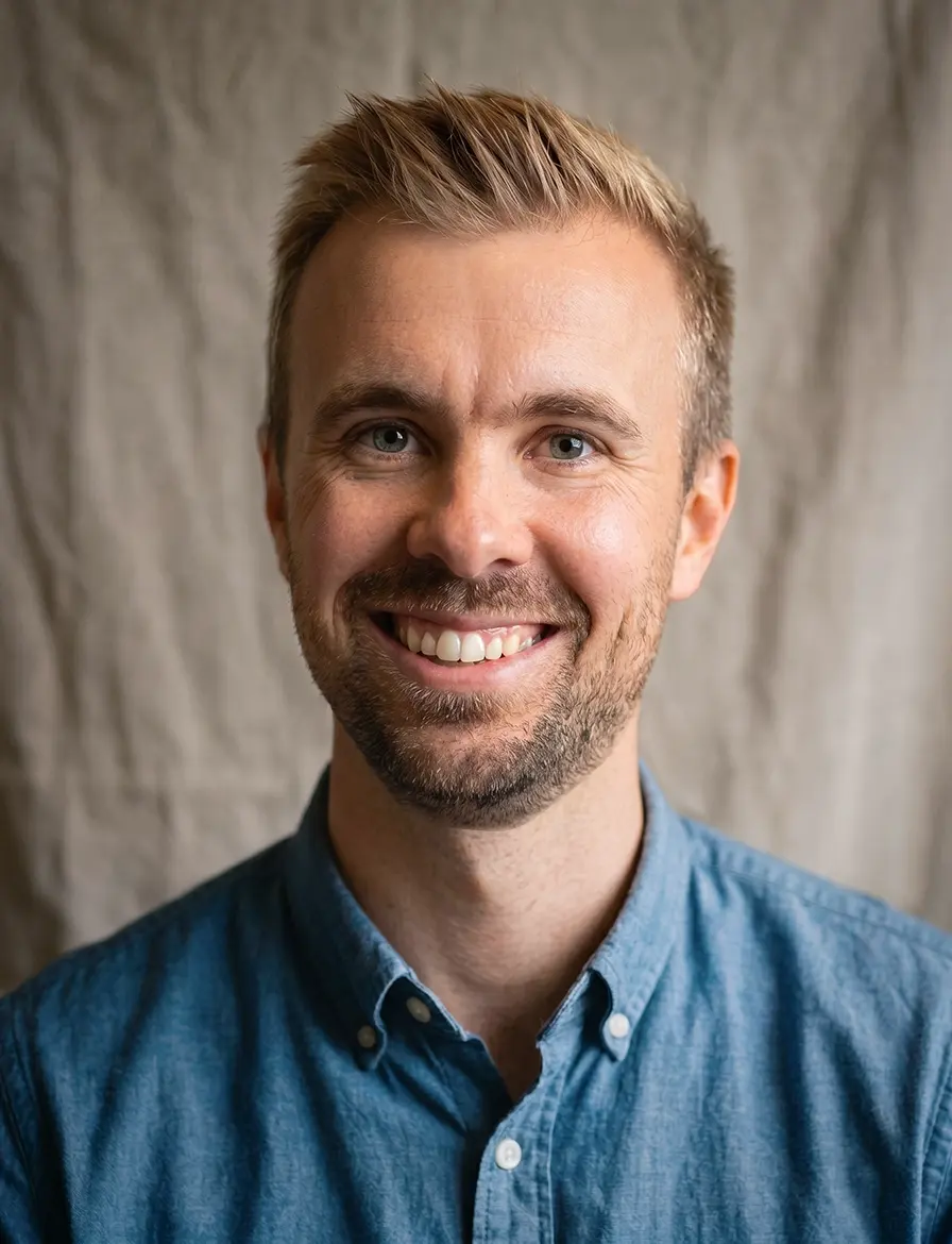Smiling man with short blond hair and beard wearing a blue button-up shirt against a plain backdrop.