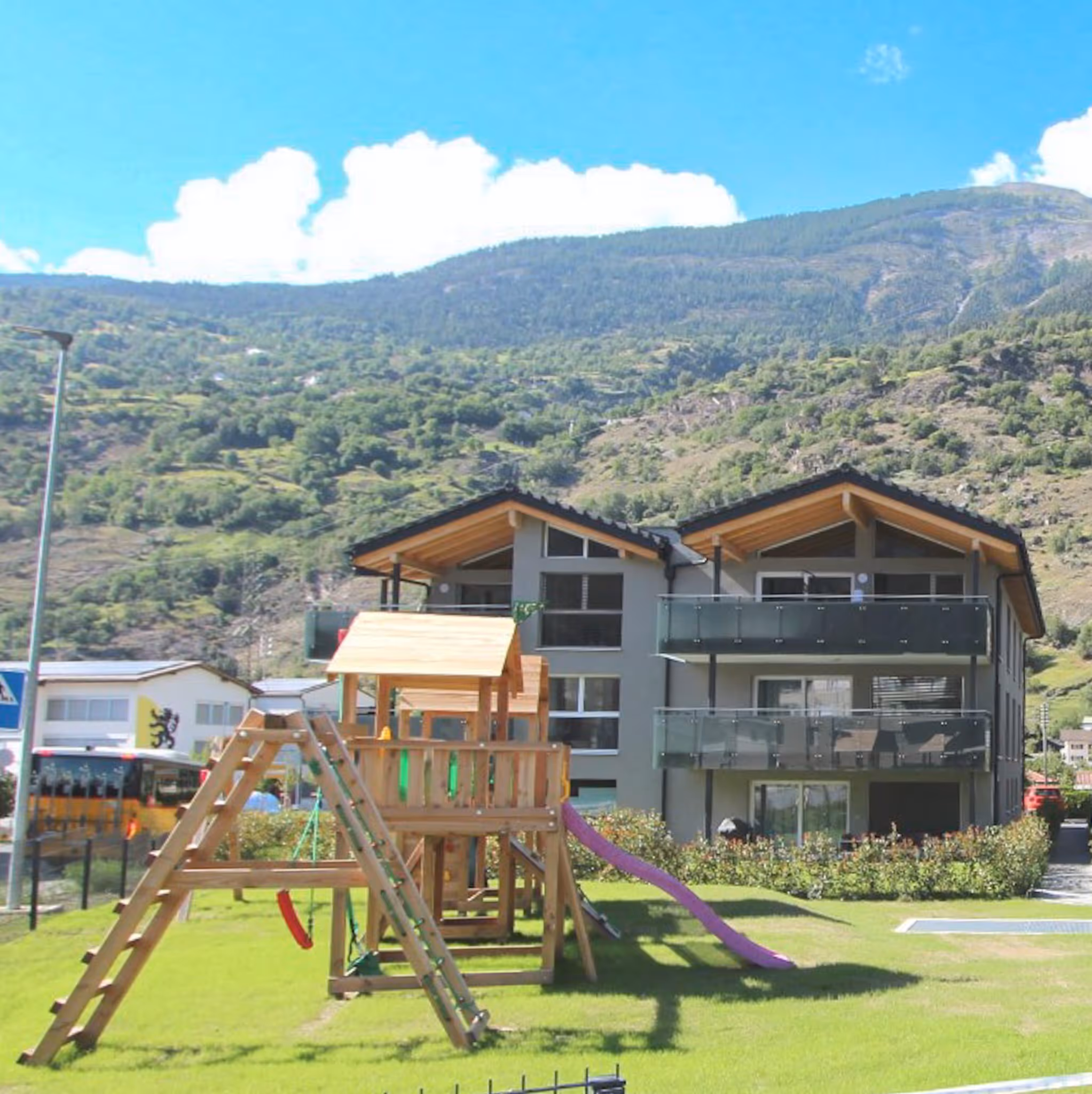 Holzspielplatz mit Rutsche vor modernen Häusern und Bergen im sonnigen Tal.