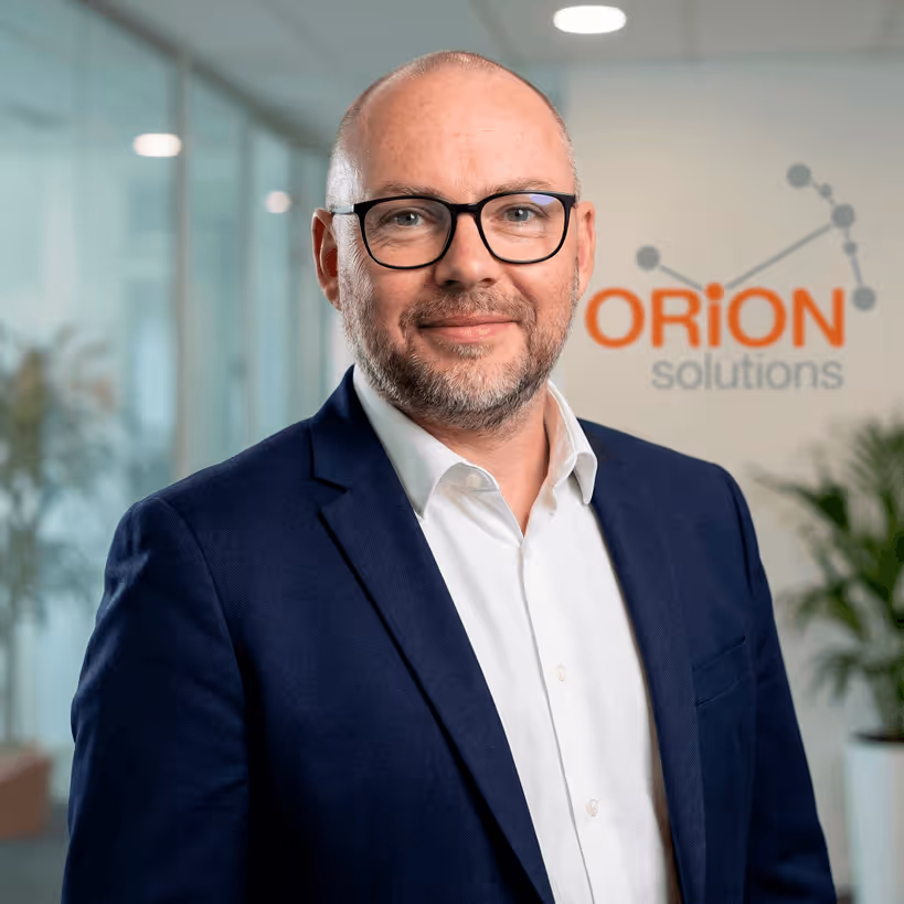 Homme chauve portant des lunettes, costume bleu et chemise blanche souriant devant un mur avec le logo 'ORiON solutions'.