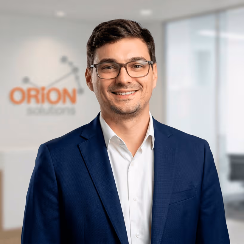 Homme souriant portant des lunettes, veste bleu marine et chemise blanche, devant un mur flou avec le logo Orion Diagnostics.