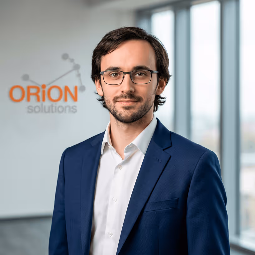 Homme en veste bleue et chemise blanche portant des lunettes, debout devant un mur avec un logo orange et gris 'ORION solutions'.
