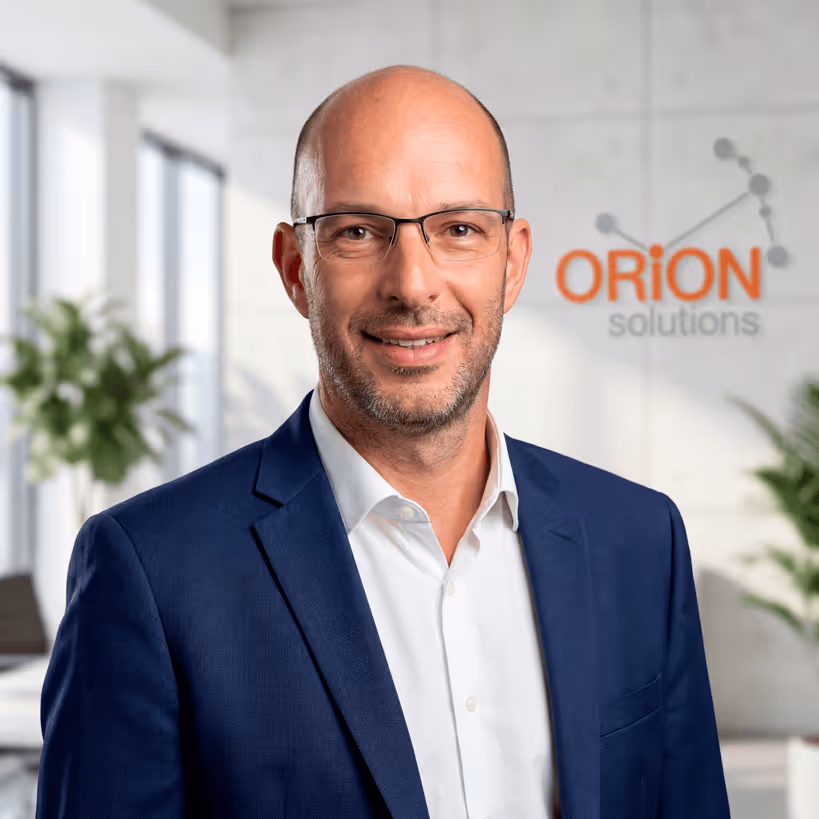Homme chauve portant des lunettes et un costume bleu souriant dans un bureau avec le logo Orion Solutions en arrière-plan.