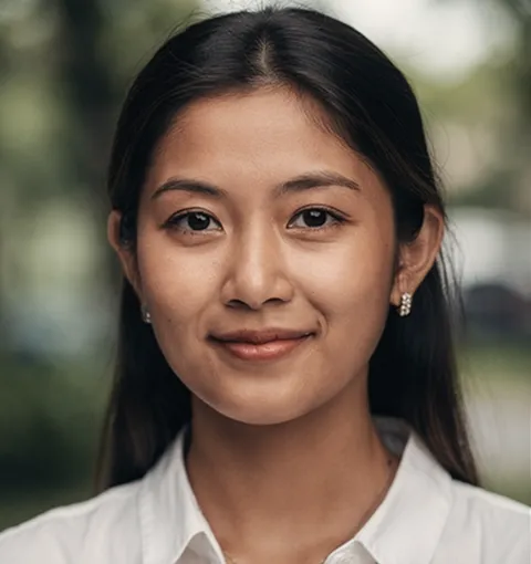 Portrait of a young woman with long dark hair, wearing a white shirt and small earrings, smiling softly with a blurred outdoor background.