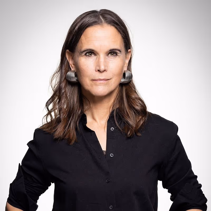 Professional headshot of Emma Ihre against a light grey background, wearing a black shirt and silver hoop earrings