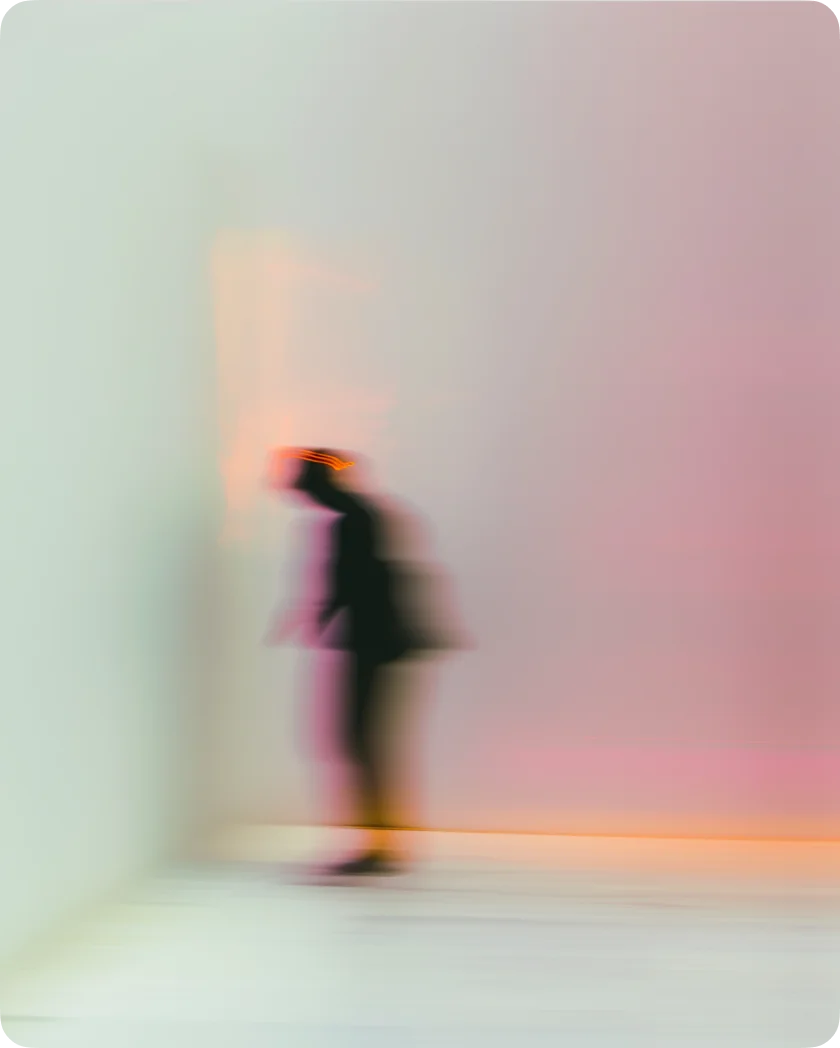 Blurry silhouette of a person standing and leaning against a wall with muted pink and orange lighting in the background.