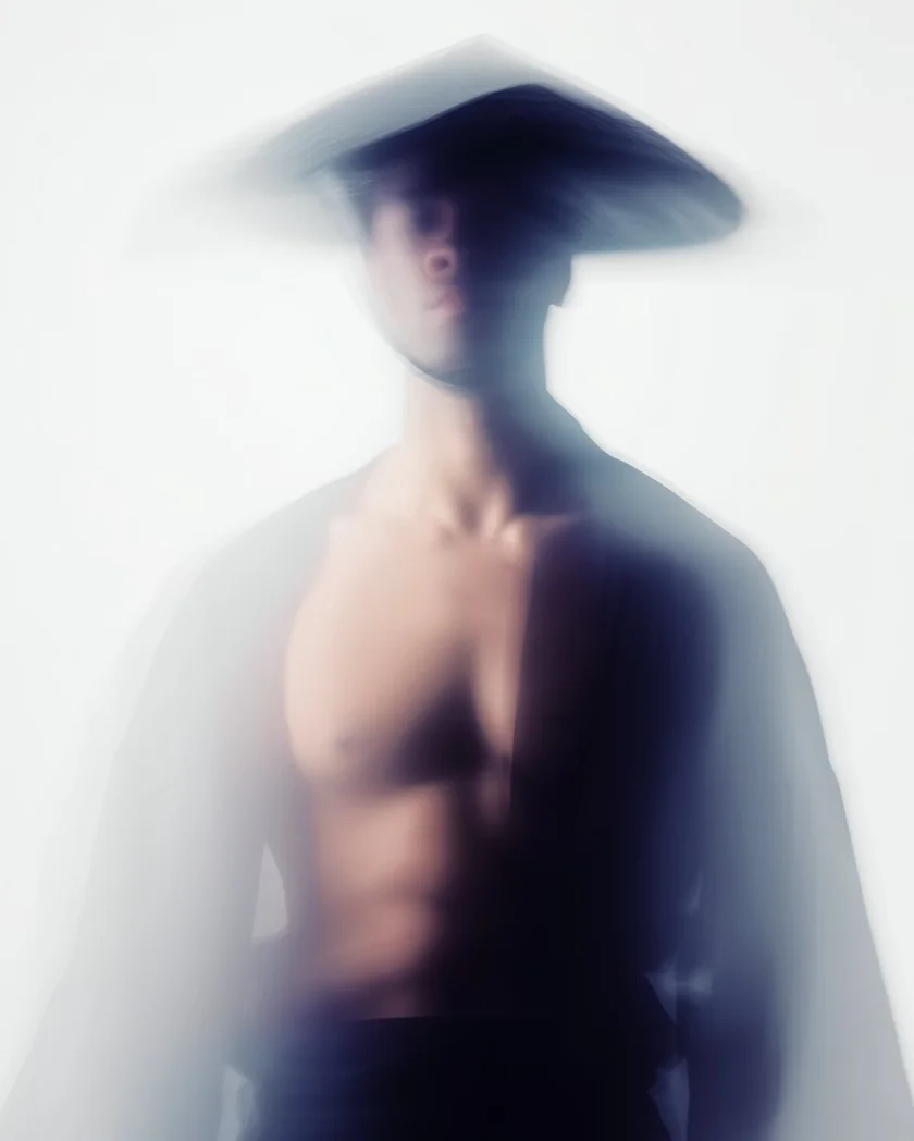 Blurred artistic portrait of a shirtless person wearing a traditional wide-brimmed conical hat against a light background.