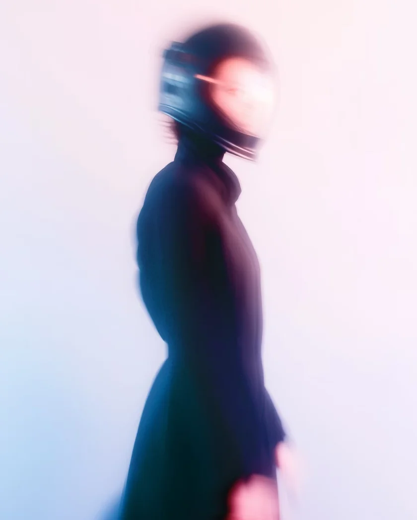 Blurred side view of a person wearing a black motorcycle helmet and black outfit against a pale background.