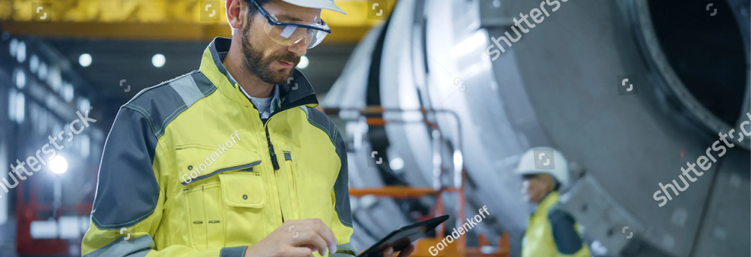 Industrial worker wearing safety gear and using a tablet inside a factory environment with heavy machinery.
