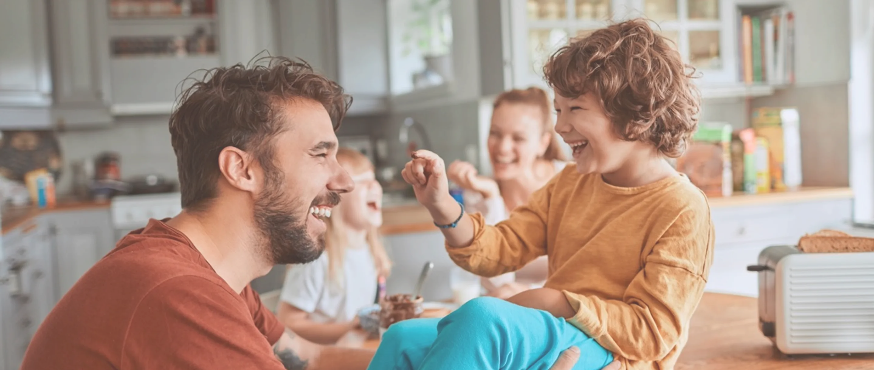 Smiling man with beard playfully interacting with a happy child sitting on a kitchen counter, with two laughing children in the background.