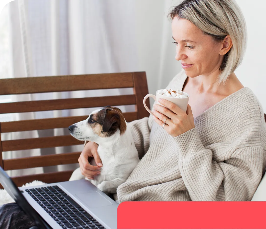 Woman in a beige sweater holding a mug of hot chocolate with marshmallows, sitting with a laptop and a small dog on her lap.