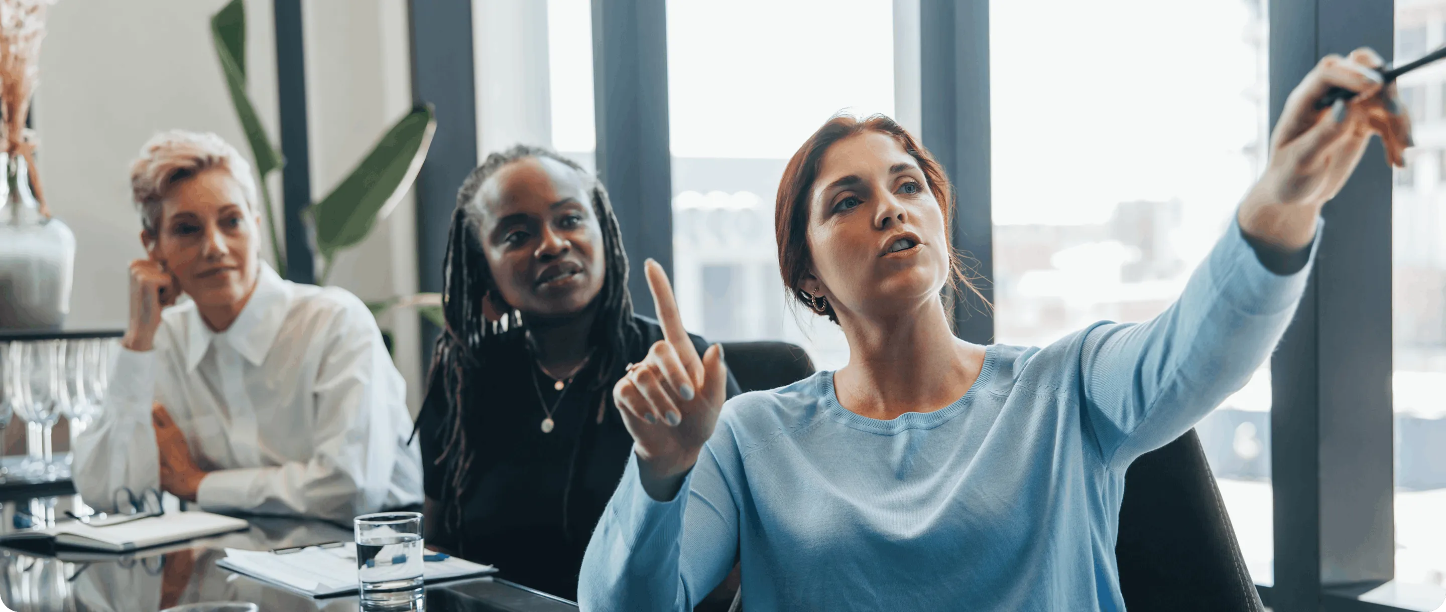 Three women in a meeting room, one in a blue sweater pointing at something outside the frame while the other two listen attentively.