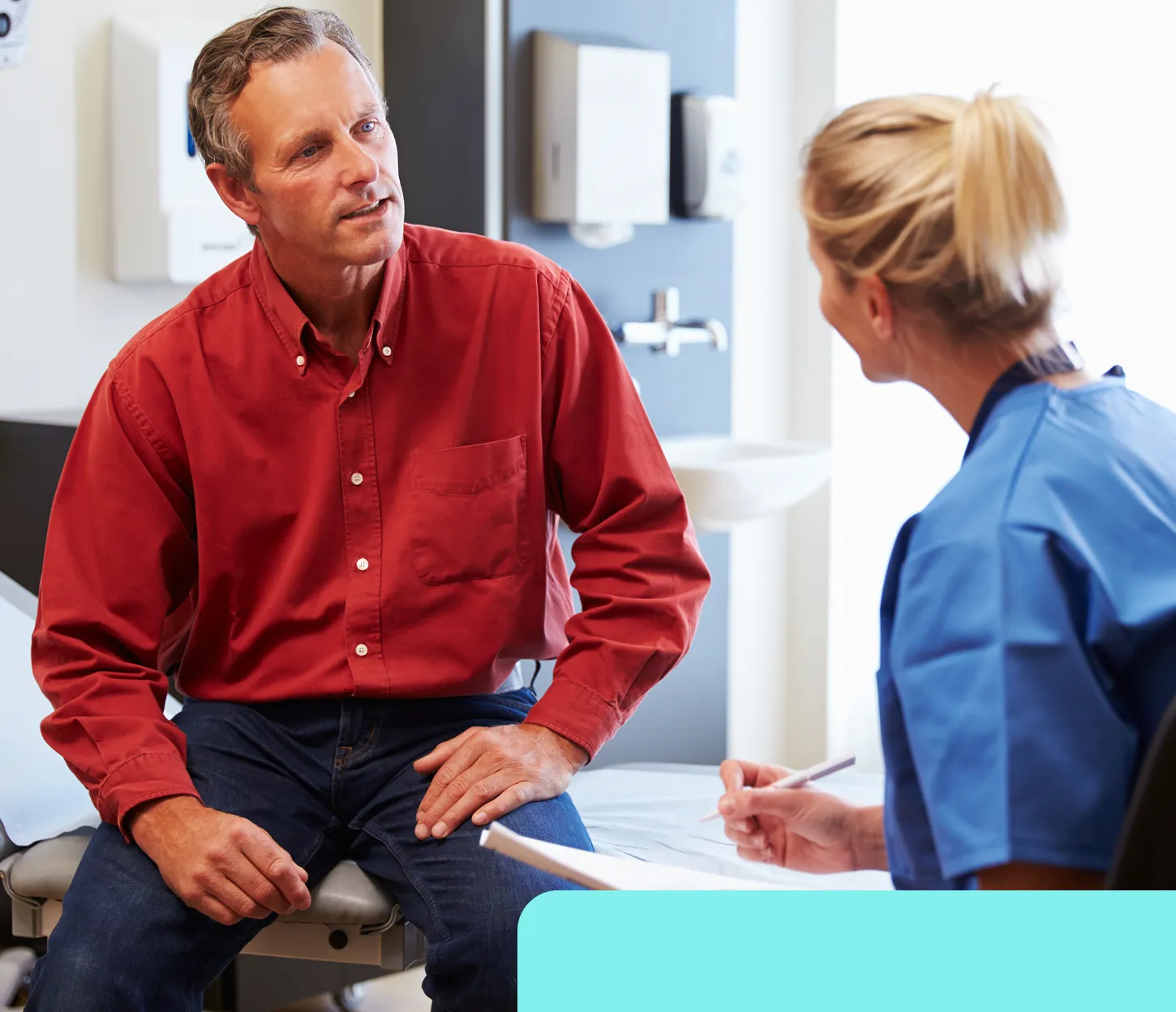 Male patient in a red shirt talking to a female healthcare provider in blue scrubs in a medical exam room.