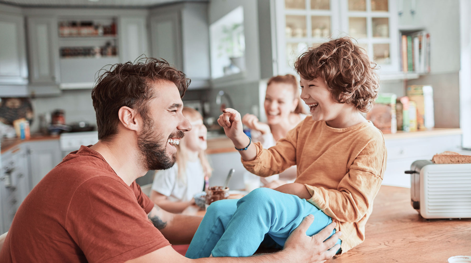 Smiling father playing with young son sitting on kitchen counter while two daughters laugh in the background.