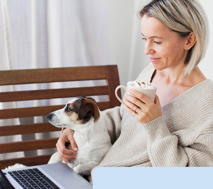 Woman in a beige sweater holding a mug of hot chocolate with marshmallows, sitting with a laptop and a small dog on her lap.