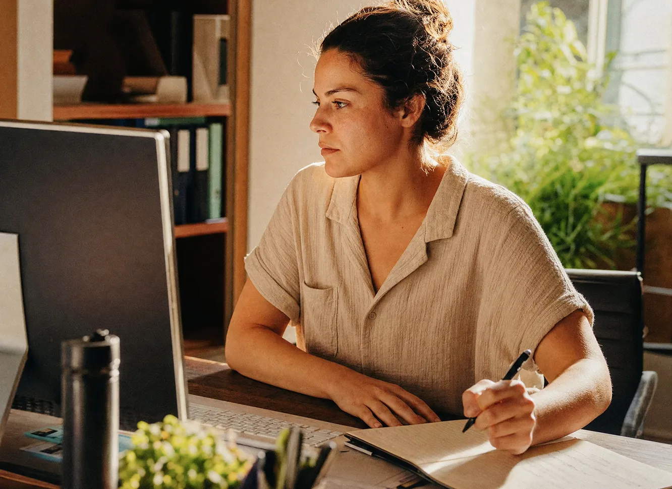 A woman wearing a light beige shirt sits at a desk in a sunlit room, looking at a computer monitor while holding a pen and writing in a notebook.