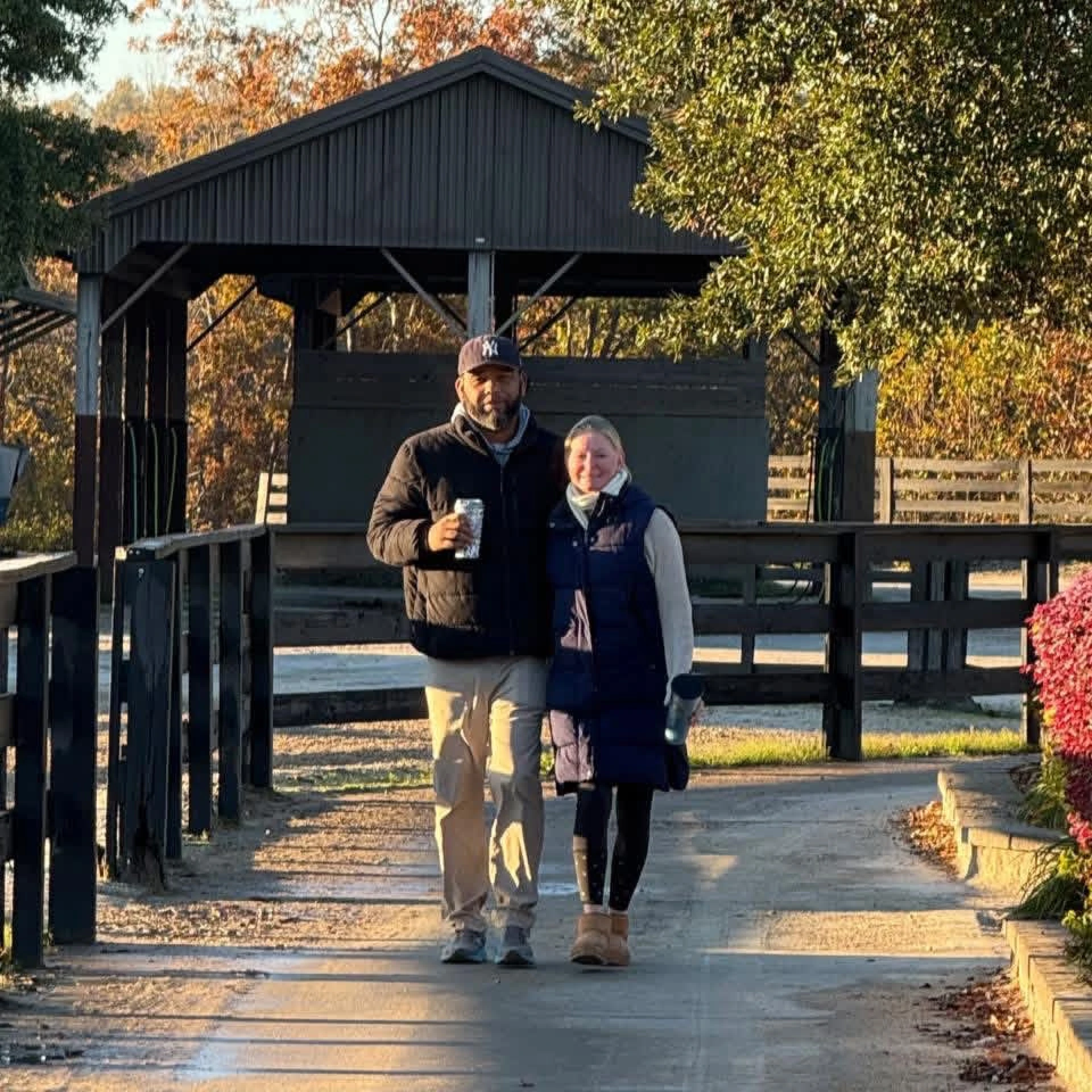Man and woman walking arm in arm on a sunlit path beside a black wooden fence and a small pavilion.