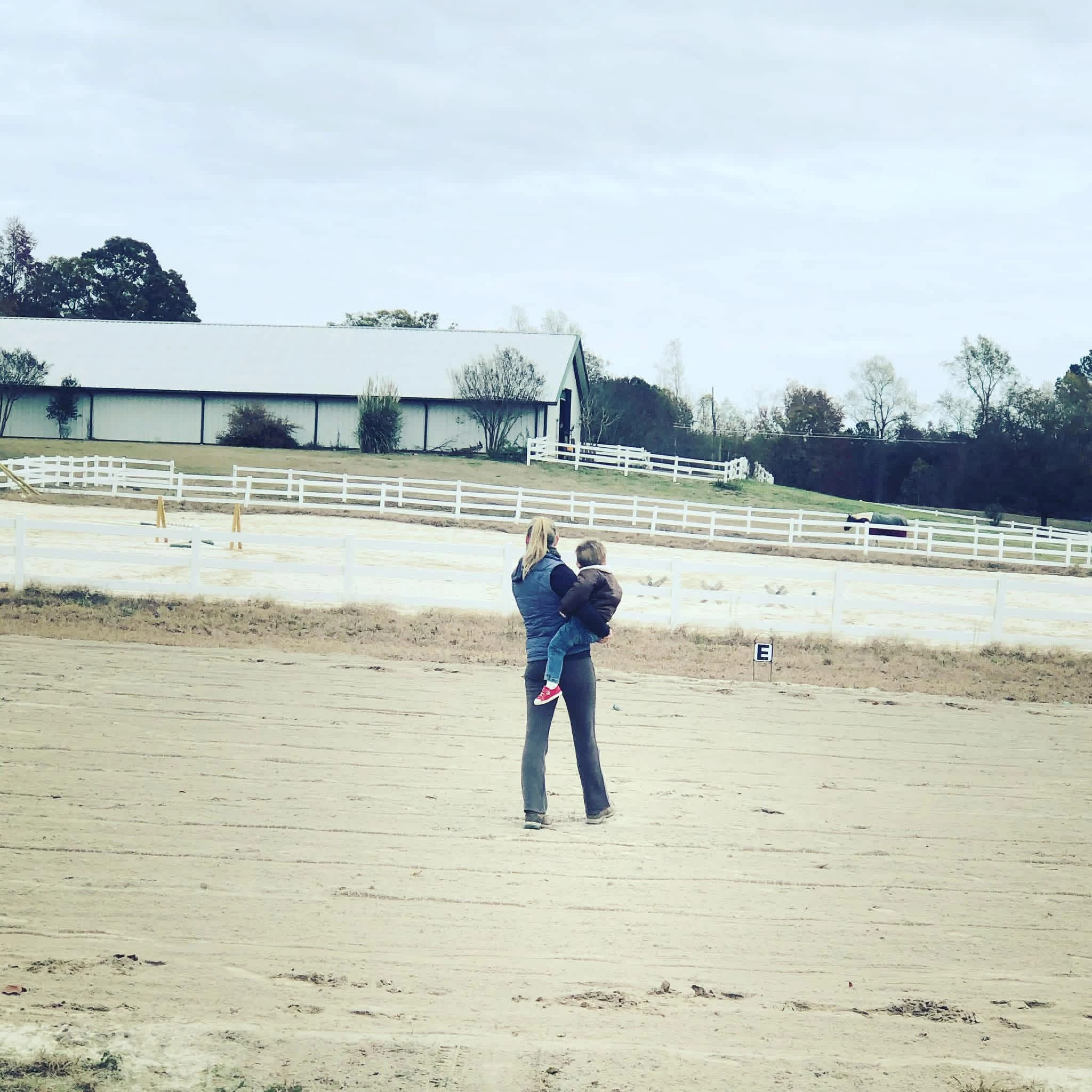 Woman holding a child walking on a sandy area near white fences with a barn and horse in the background.