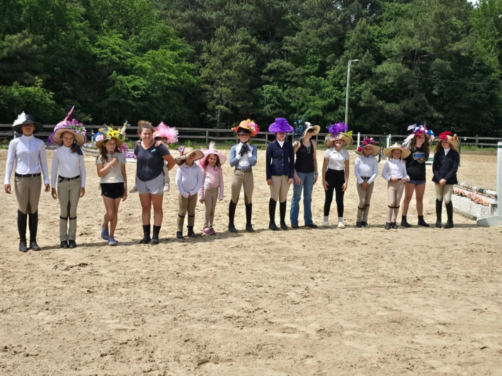 Group of children and a woman standing in a sandy outdoor area, wearing decorative hats and a mix of riding attire and casual clothing, with trees in the background.