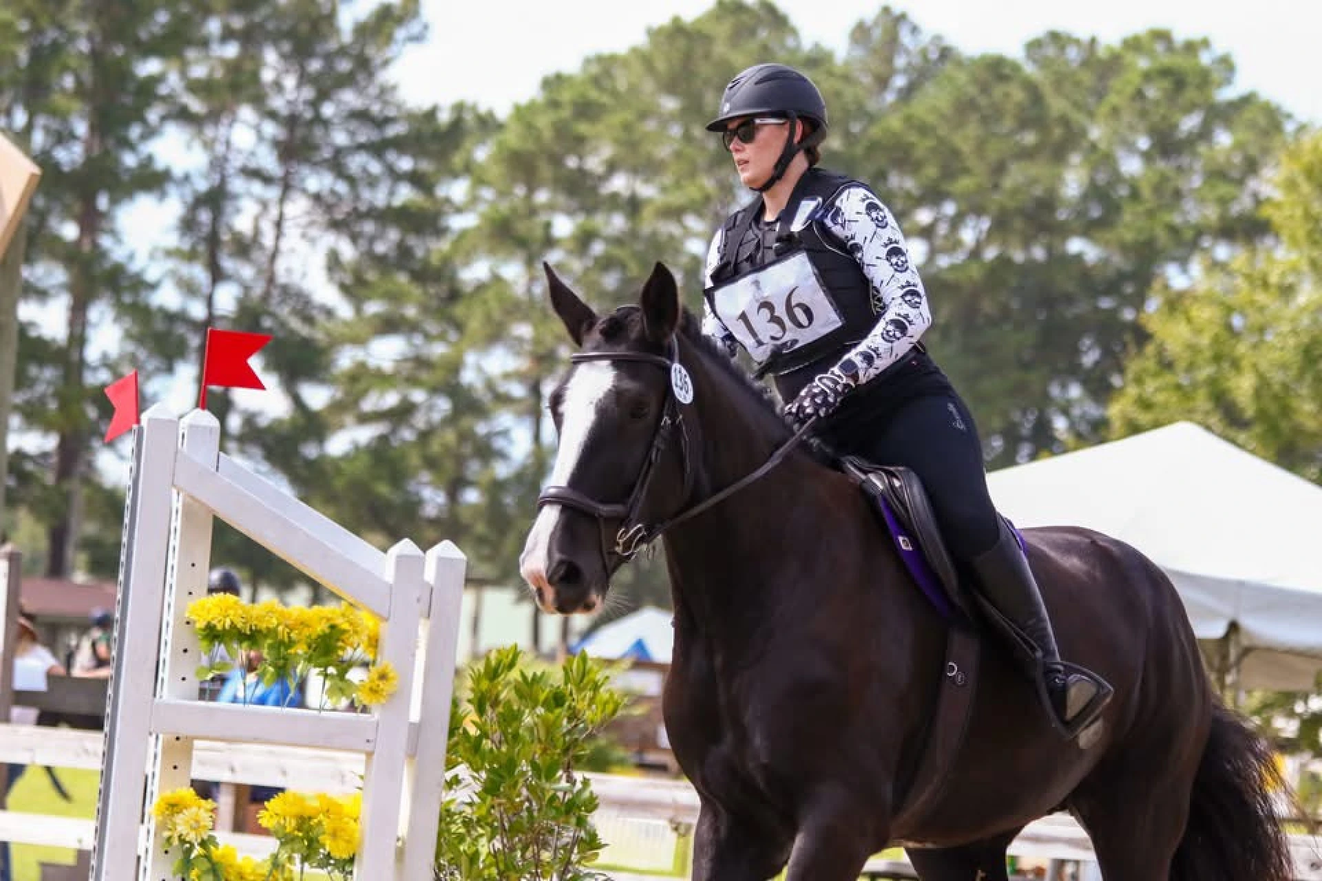 Equestrian rider in helmet and protective vest on a dark horse during a show jumping event near a white fence decorated with yellow flowers and red flags.