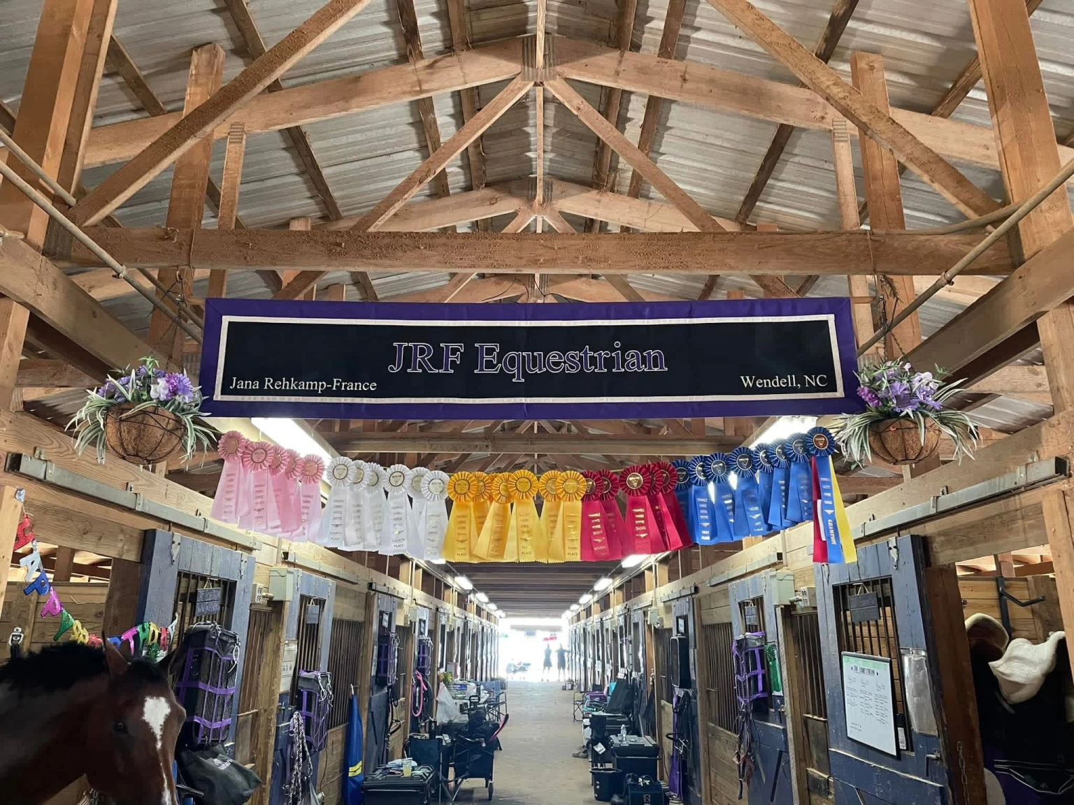 Interior of a wooden horse stable with a JRF Equestrian banner and rows of colorful ribbons hanging above horse stalls.