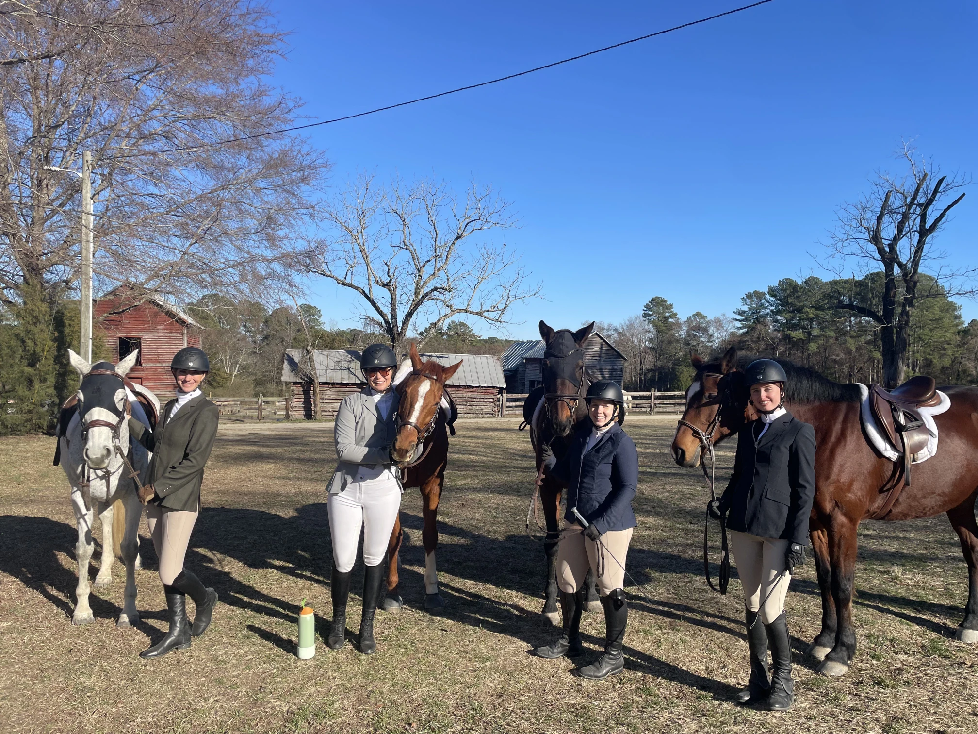 Four women in equestrian attire standing outdoors with their horses in front of wooden barns under a clear blue sky.