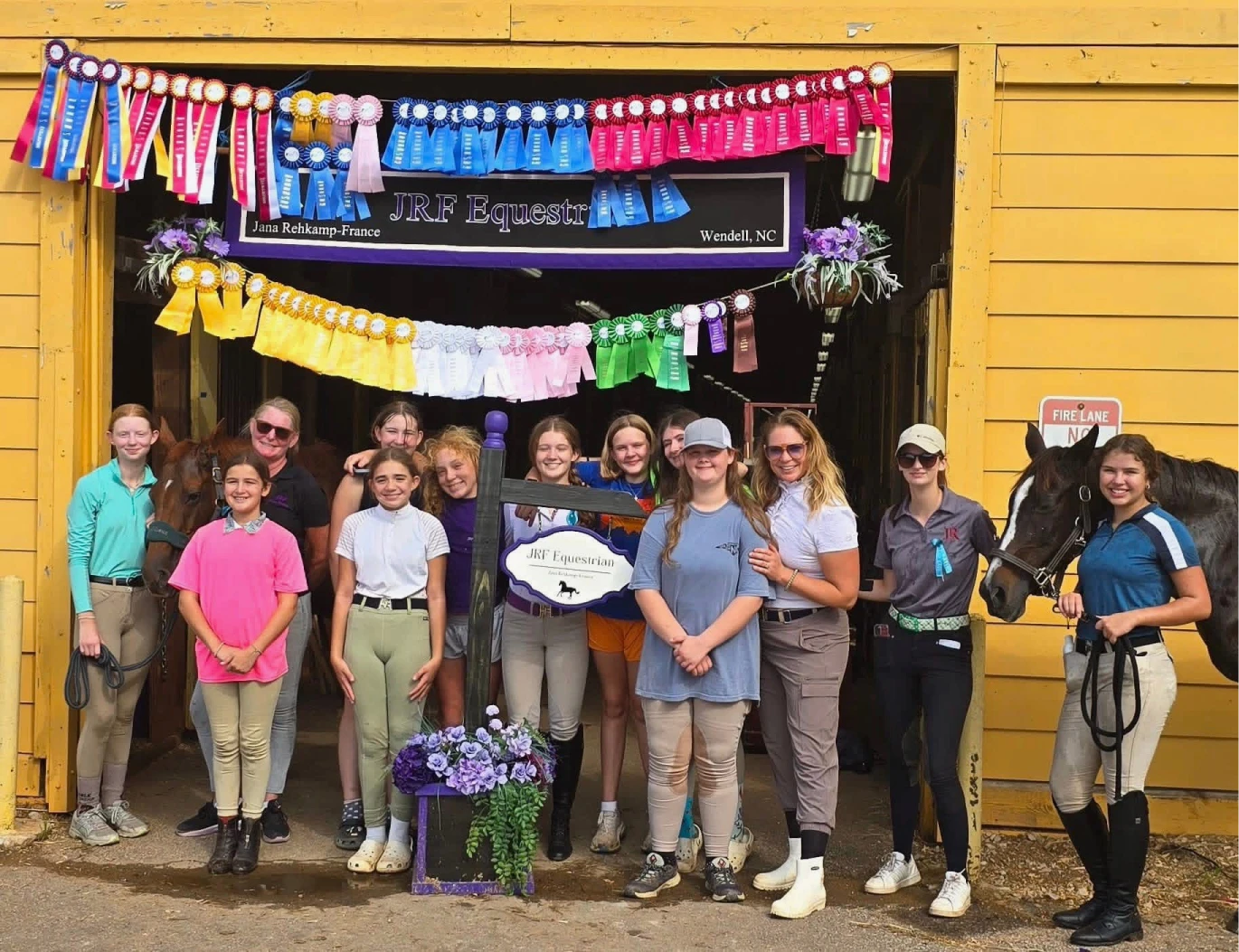 Group of young equestrians and two adults standing in front of a barn with colorful horse show ribbons displayed above and two horses on each end.