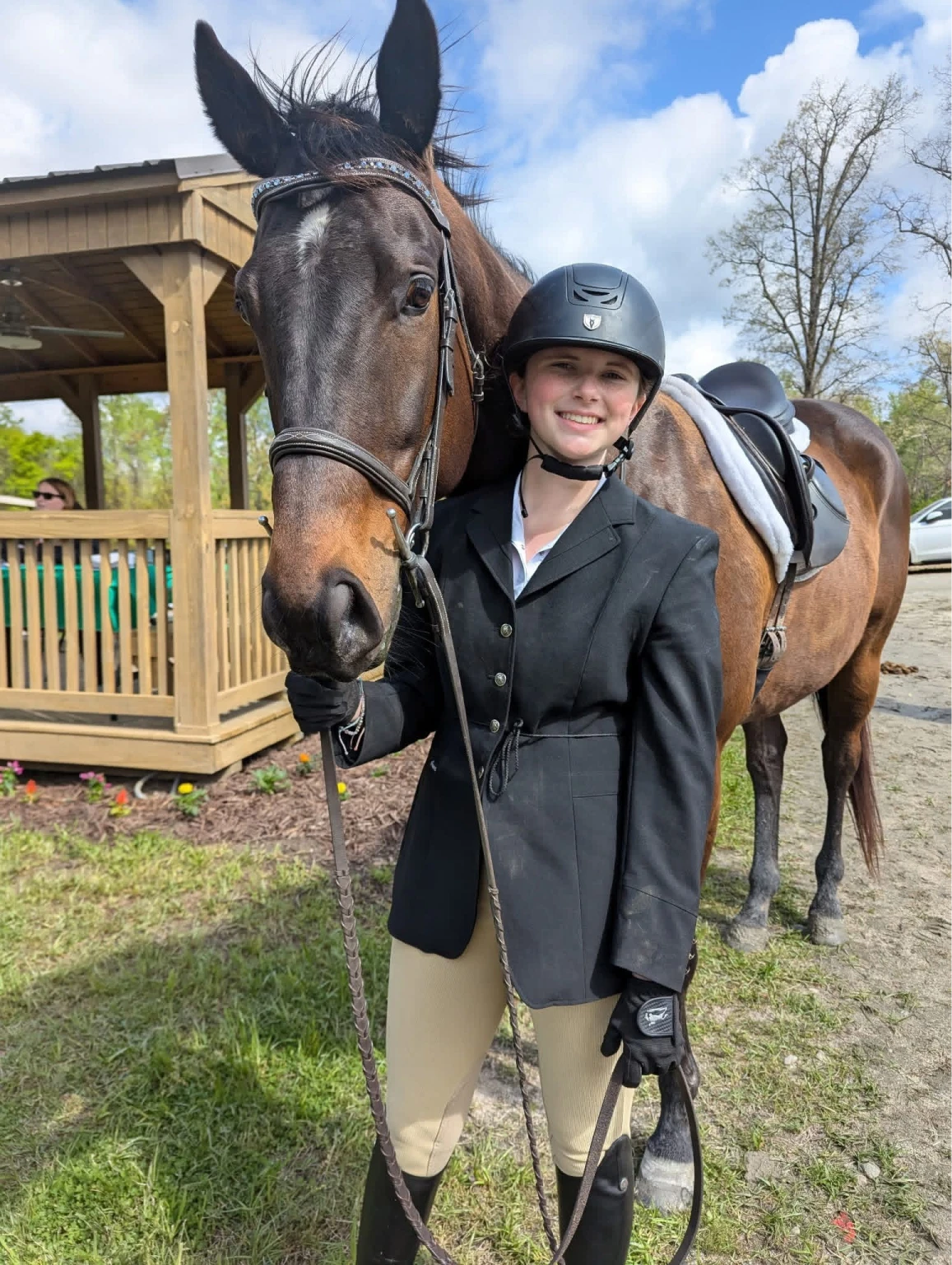 Smiling young equestrian in riding helmet and jacket standing next to a saddled brown horse outdoors.