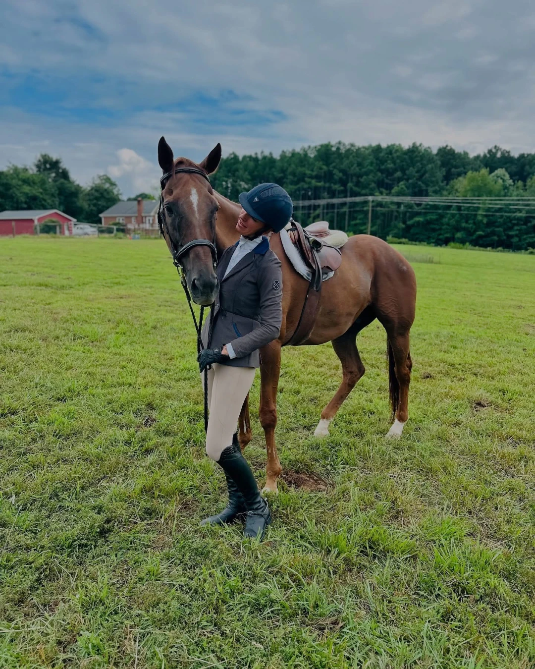 Person in equestrian attire standing on grass field, affectionately embracing a saddled brown horse.