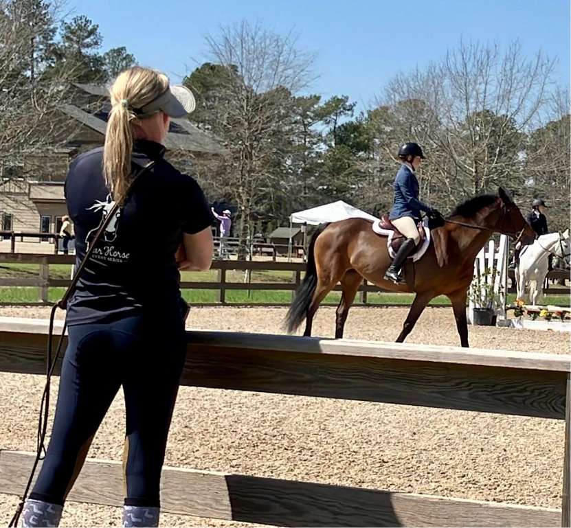 Person in black watching a rider on a brown horse in an outdoor equestrian arena on a sunny day.